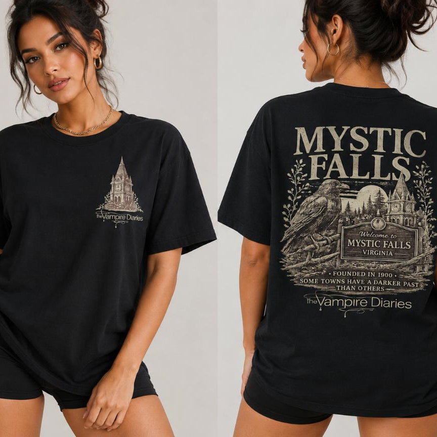 A woman modeling a black short-sleeve graphic T-shirt shown from both front and back against a light gray studio background. The front has a small white line-art graphic on the left chest, and the back has a large white graphic reading 'MYSTIC FALLS' with additional decorative text and illustration.